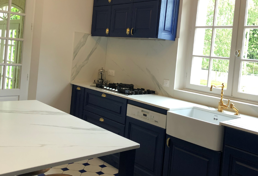 Évier blanc à robinet doré dans une cuisine bleu marine, avec îlot à effet marbre, pour Arboucave