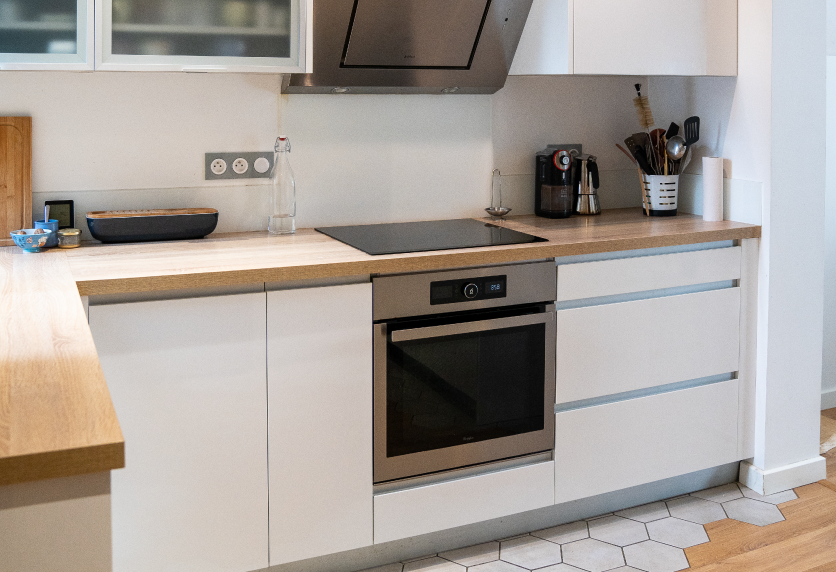 Pour une maison à Audignon, cuisine blanche avec plan de travail en bois, plaque de cuisson et four encastré