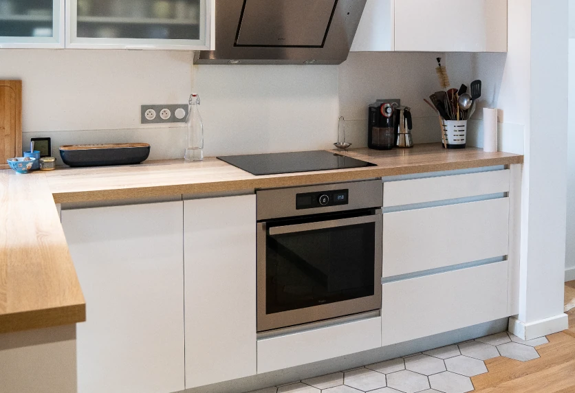 Pour une maison à Audignon, cuisine blanche avec plan de travail en bois, plaque de cuisson et four encastré
