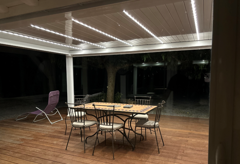 À Audignon, pergola blanche avec éclairage intégré au-dessus d’une table installée sur la terrasse en bois.