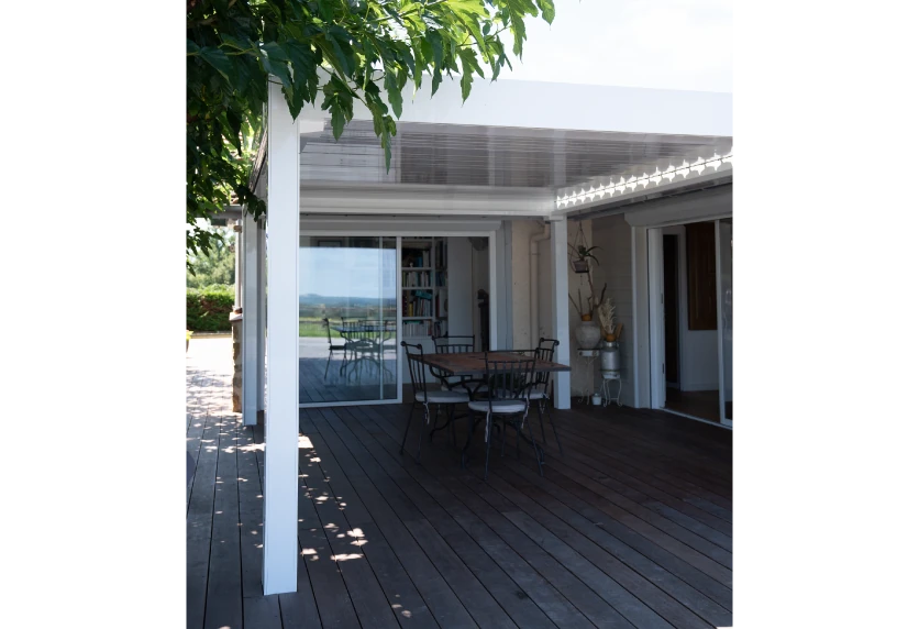 Sur une terrasse en bois à Samadet, une pergola blanche abrite une table devant les baies vitrées.