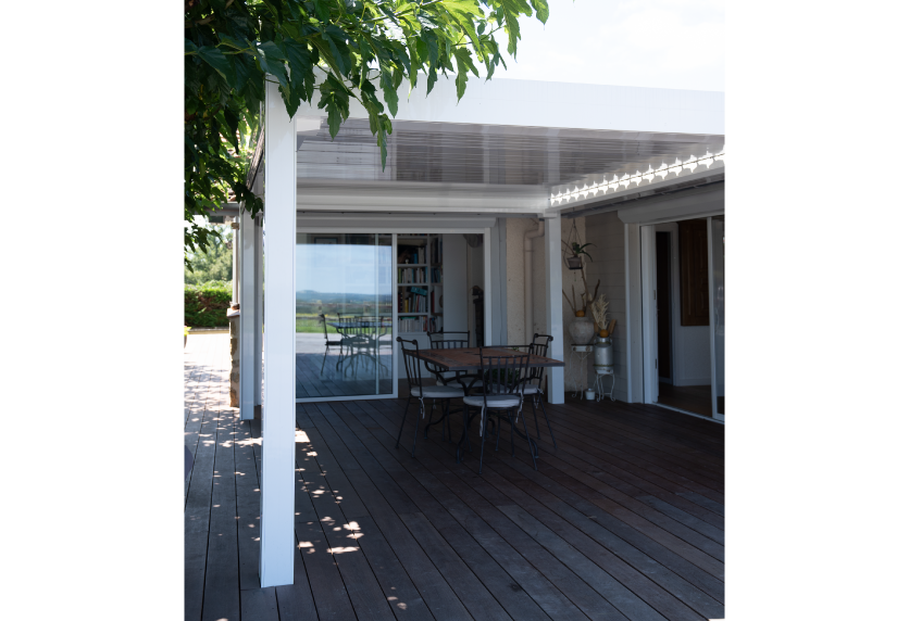 Sur une terrasse en bois à Samadet, une pergola blanche abrite une table devant les baies vitrées.