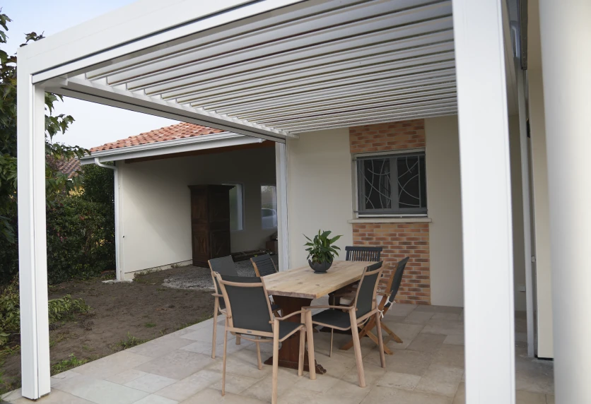 À Geaune, une pergola blanche à lames surplombe la table et les chaises disposées devant la maison.
