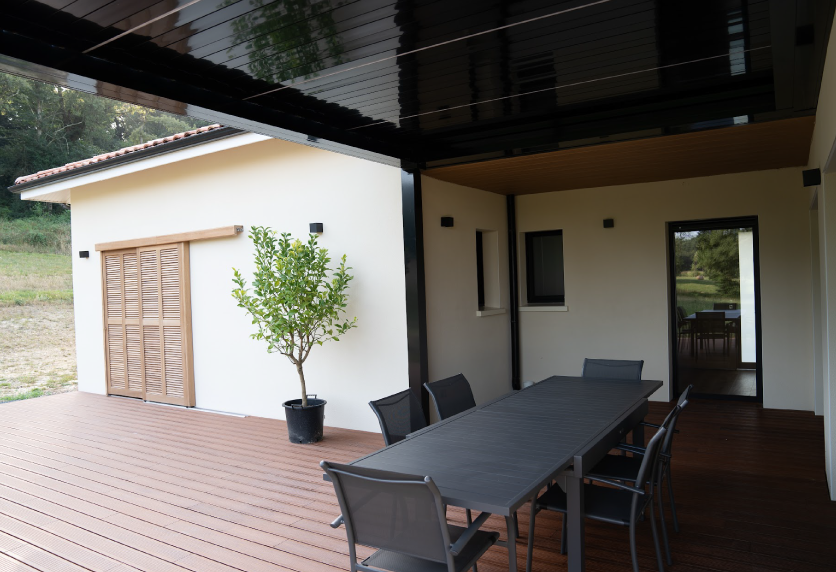 Depuis la table de terrasse, vue sous une pergola foncée adossée à la maison, pour un projet à Pimbo.