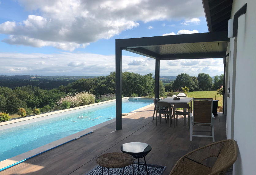Une pergola sombre protège le coin repas sur une terrasse en bois bordant la piscine, dans ce projet de menuiserie à Miramont-Sensacq.
