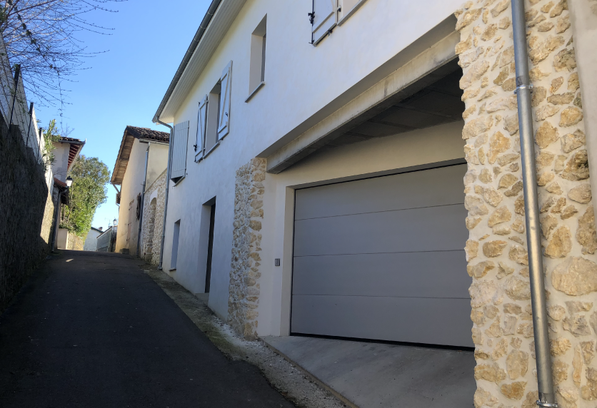 Dans une rue de Samadet, une porte de garage gris clair ferme le rez-de-chaussée d’une maison aux murs de pierre