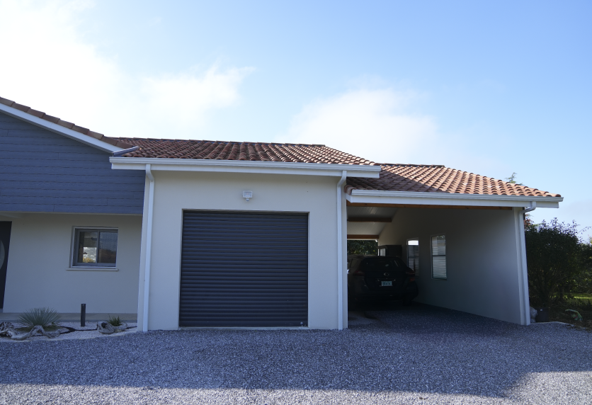 Pour une maison à Castelnau-Tursan, porte de garage grise attenante à un abri couvert sur une façade blanche.