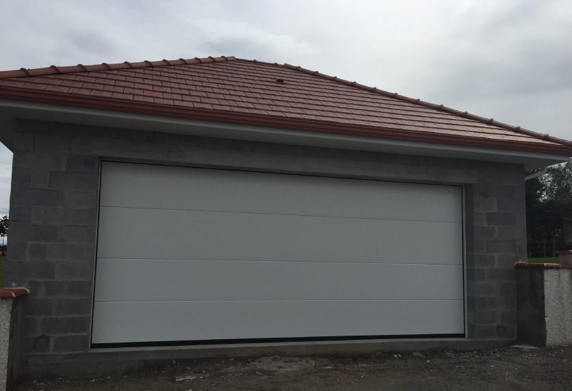 Large porte de garage blanche installée sur un bâtiment aux murs gris et au toit de tuiles, pour un projet à Clèdes.