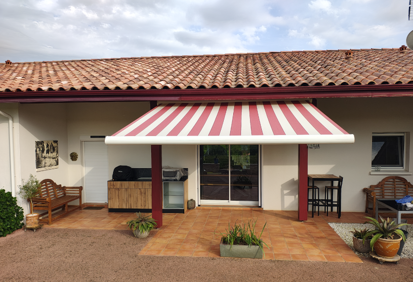 Sous un toit en tuiles, store banne rouge et blanc ombrageant une terrasse, dans un projet de menuiserie extérieure vers Garlin